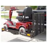 A red mobility scooter is securely mounted on a black Versahaul carrier attached to the back of a black pickup truck. The scooter has a basket in front, a cushioned seat, and large wheels. The setup is parked in a parking lot.