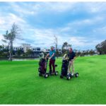Two individuals joyfully riding the Golf Skate Caddy GSC™ Tourer X Golf Cart Mobility Scooter across a golf course, with a tranquil water lake in the background, adding to the picturesque scene of outdoor recreation.