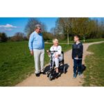 A woman on an eFOLDi Lite scooter engaged in conversation with a man and a young boy during a sunny day in the park.