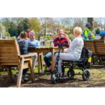 A woman seated on an eFOLDi Lite scooter at an outdoor table with a family, in a park-like setting.