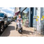A woman riding an eFOLDi Lite scooter on a street, with shops and parked cars around.