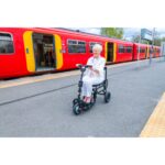 A woman seated on an eFOLDi Lite scooter at a train station platform, with a red train in the background.