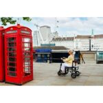 A senior woman riding the eFOLDi Lite scooter near a traditional red telephone box, demonstrating the scooter's maneuverability and practicality for sightseeing and city navigation.