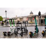 The eFOLDi Lite scooter parked amongst public rental scooters in a city square, showcasing its compact design and readiness for urban mobility next to other transport options.