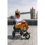 A user operating the eFOLDi Explorer mobility scooter on a city promenade, with a view of the river and a historic bridge, highlighting the scooter's functionality in touristic locations.