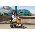 A user riding the eFOLDi Explorer mobility scooter across a bridge with the cityscape in the background, displaying the scooter's use in traversing public spaces.
