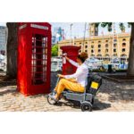 A person sitting on the eFOLDi Explorer scooter by a red telephone box and mail pillar box, illustrating the scooter in use in an urban outdoor setting.