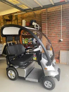 A stylish, enclosed mobility scooter with a black seat, armrests, and a front windshield for weather protection.