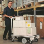 Man in work uniform using EK Tech Pony Express 1031 motorized platform cart loaded with cardboard boxes in a warehouse aisle